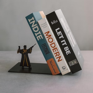 Könnte beinhalten: Ein schwarzer Metall-Buchstütze mit einer Silhouette eines Zauberers, der einen Zauberstab hält und drei Bücher stützt. Die Bücher tragen die Titel "Indie Festival", "Modern" und "Let It Be". Die Buchstütze steht auf einer grauen Oberfläche.