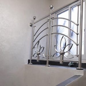 Puede incluir: Una barandilla de acero inoxidable pulido con diseños florales y postes superiores redondeados. La barandilla está instalada en una escalera, con una ventana visible en el fondo. El diseño incorpora barras curvas y elementos florales decorativos.