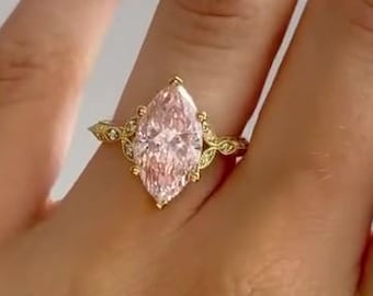 May include: A gold ring with a light pink, marquise-cut gemstone. The ring has an intricate design with small, clear accent stones. The ring is set on a hand with pink nails.