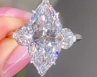 Könnte beinhalten: Ein Diamantring mit einem großen, birnenförmigen Mittelstein, flankiert von kleineren, runden Diamanten. Das Design des Rings zeigt eine Ansammlung von Brillanten, die einen funkelnden Effekt erzeugt. Der Ring wird von einer Person gehalten.