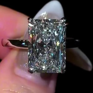 May include: A platinum ring featuring a large, rectangular radiant-cut diamond. The diamond's facets catch the light, creating a dazzling display. The ring is set against a black backdrop, accentuating the diamond's sparkle. A timeless piece of jewellery.