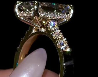 Può includere: Un anello d'oro con una grande pietra preziosa a taglio rettangolare come elemento centrale. L'anello presenta pietre più piccole lungo la fascia e intorno alla pietra principale, il tutto su sfondo nero. L'anello è tenuto dalle dita di una persona.