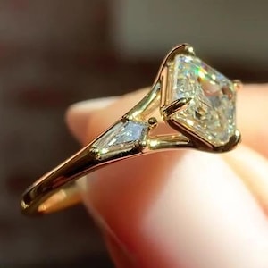 May include: A gold ring with a large, rectangular-cut diamond as the focal point. Two smaller, baguette-cut diamonds are set on either side. The ring's band is polished gold, presenting a classic and elegant design.