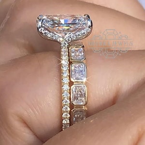 May include: Close-up of a gold ring featuring a large, rectangular-cut diamond, encircled by smaller diamonds. A second gold band with rectangular diamonds is also visible. The rings are on a hand.
