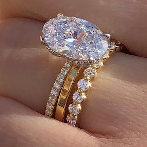 May include: A gold engagement ring featuring a large oval-cut diamond, surrounded by smaller diamonds on the setting. Two additional gold bands with diamond detailing are stacked with the engagement ring.
