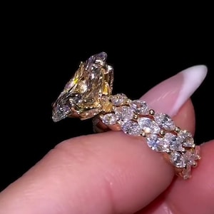 May include: A gold ring featuring a large, irregularly shaped yellow gemstone and a band set with numerous small, clear gemstones. The ring is displayed against a black backdrop, accentuating its sparkle and detailed design. A luxurious piece of jewellery.