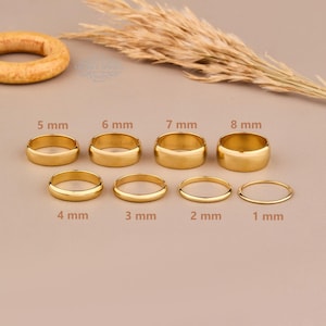 Może przedstawiać: Kolekcja złotych pierścionków o różnych szerokościach, od 1 mm do 8 mm. Rozmiary są wyraźnie oznaczone. W tle element dekoracyjny.