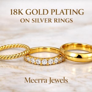 May include: Three gold-plated silver rings are displayed on a marble surface. The rings include a twisted band, a band with clear stones, and a simple band. Text reads "18K GOLD PLATING ON SILVER RINGS" and "Meerra Jewels."