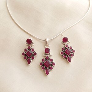 May include: A silver jewellery set consisting of a necklace with a pendant and two matching drop earrings. Each piece features a central square-cut deep pink gemstone surrounded by smaller round and teardrop-shaped pink stones in silver settings.