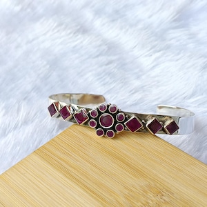 Può includere: Un bracciale a polsino in argento con un design floreale e pietre preziose di rubino taglio quadrato. Il bracciale presenta un fiore centrale con un grande rubino circondato da rubini più piccoli. Il bracciale è esposto su una superficie di legno.