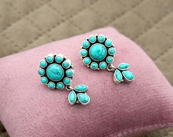 May include: A pair of silver earrings with turquoise stone detailing. Each earring has a central turquoise stone encircled by smaller stones, with a cluster of three turquoise stones hanging below. The earrings are presented on a pink surface.