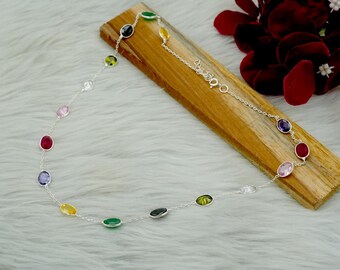 May include: A silver necklace featuring a series of colorful, oval-shaped gemstones. The gemstones are set along a delicate chain, with a variety of colors including red, pink, yellow, green, and purple. The necklace is displayed on a wooden surface.