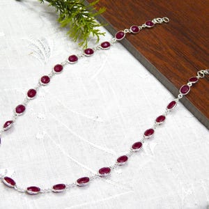 Può includere: Una collana d'argento con pietre preziose ovali di colore rosso intenso. Le pietre sono incastonate in castoni d'argento individuali e collegate tra loro. La collana è esposta su una superficie bianca con sfondo in legno.