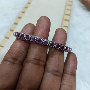 Può includere: Un braccialetto rigido argentato con una fila di pietre preziose rotonde, color magenta intenso. Le pietre sono incastonate in castoni individuali lungo il braccialetto. Il braccialetto è tenuto in mano, mostrando il suo design e colore.