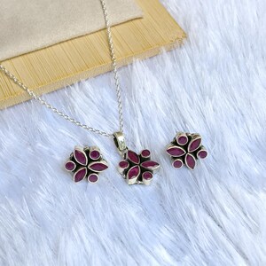 May include: A silver necklace, pendant, and stud earrings set. Each piece has a flower design with five petals and a central ruby-coloured stone. The jewellery set is displayed on a white, fluffy surface.