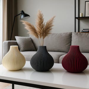 May include: Three decorative vases in cream, black, and burgundy, with a ribbed design, sit on a white table. The black vase holds dried pampas grass. The vases are in a living room setting.