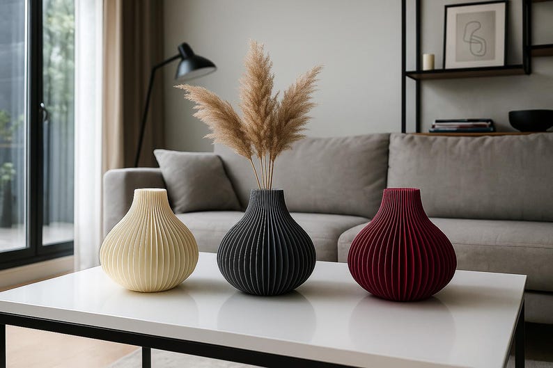 May include: Three decorative vases in cream, black, and burgundy, with a ribbed design, sit on a white table. The black vase holds dried pampas grass. The vases are in a living room setting.