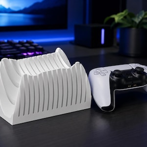 Op de afbeelding: Een witte plastic gaming controller standaard met een reeks verticale, gebogen vinnen ontworpen om een controller vast te houden. Naast de standaard staat een witte en zwarte draadloze gaming controller met twee analoge sticks en knoppen.