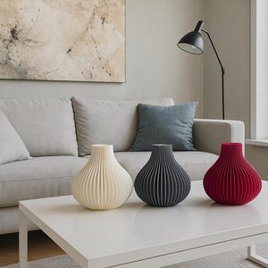 May include: Three decorative vases in cream, charcoal grey, and red sit on a white table. The vases have a ribbed design and a bulbous shape. The background includes a grey sofa, a blue cushion, and abstract art.