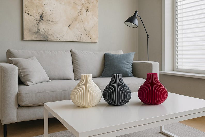 May include: Three decorative vases in cream, charcoal grey, and red sit on a white table. The vases have a ribbed design and a bulbous shape. The background includes a grey sofa, a blue cushion, and abstract art.