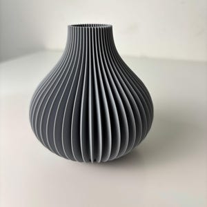 May include: A grey vase with a ribbed design. The vase has a bulbous body and a narrow neck. The vertical ribs create a textured surface. The vase is made of a solid material and is suitable for home decor.