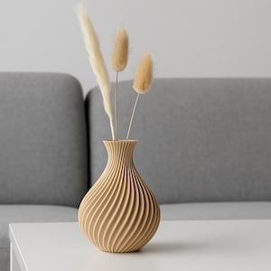 Könnte beinhalten: Eine beige Vase mit Spiralmuster, gefüllt mit getrockneten Blumen. Die Vase steht auf einem weißen Tisch, im Hintergrund ein graues Sofa. Die Vase hat ein strukturiertes Muster und ist mit getrockneten Gräsern gefüllt.