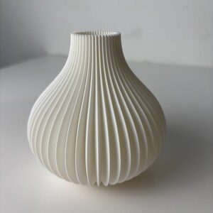 May include: A white vase with a rounded body and a slender neck. The vase has a ribbed, vertical pattern, giving it a textured appearance. The vase is made of a solid, opaque material and is suitable for home decoration.