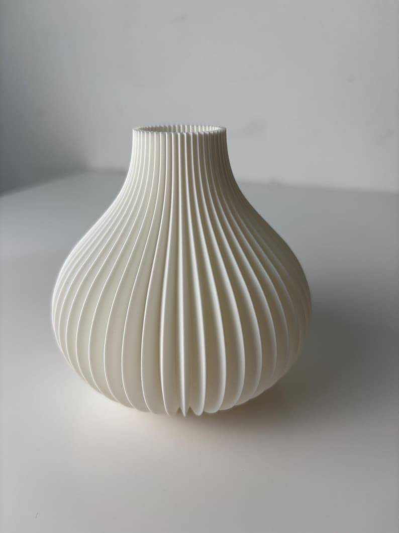 May include: A white vase with a rounded body and a slender neck. The vase has a ribbed, vertical pattern, giving it a textured appearance. The vase is made of a solid, opaque material and is suitable for home decoration.