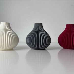 May include: Three decorative vases in a row, each with a unique colour and design. The vases are white, dark grey, and red, and have a ribbed, onion-like shape. The vases are sitting on a white surface.