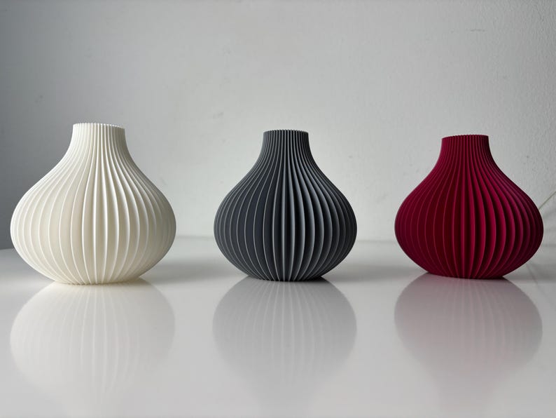 May include: Three decorative vases in a row, each with a unique colour and design. The vases are white, dark grey, and red, and have a ribbed, onion-like shape. The vases are sitting on a white surface.