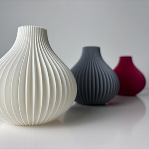May include: Three decorative vases with a ribbed design. The first is white, the second is grey, and the third is a deep red. The vases have a rounded shape and a narrow neck. The vases are arranged in a row.
