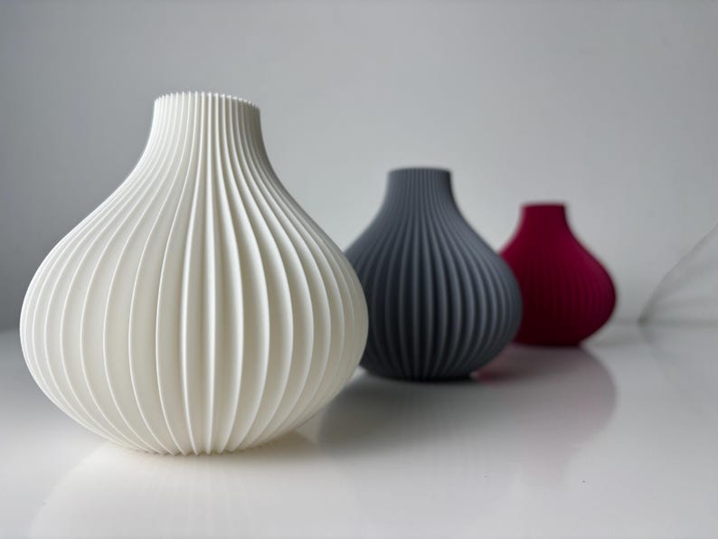 May include: Three decorative vases with a ribbed design. The first is white, the second is grey, and the third is a deep red. The vases have a rounded shape and a narrow neck. The vases are arranged in a row.