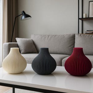 May include: Three decorative vases in cream, black, and burgundy, each with a ribbed, onion-shaped design. The vases are displayed on a white table in a living room setting. Home decor.
