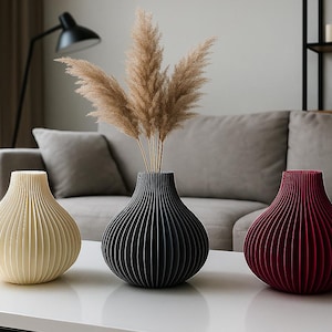 Puede incluir: Tres jarrones decorativos en crema, negro y burdeos, cada uno con un diseño acanalado. Los jarrones están colocados sobre una mesa blanca, con hierba de la pampa seca en el jarrón negro. El fondo incluye un sofá gris y una lámpara negra.