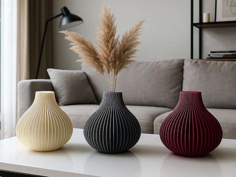 May include: Three decorative vases in cream, black, and burgundy, each with a ribbed design. The vases are arranged on a white table, with dried pampas grass in the black vase. The background includes a grey sofa and a black lamp.