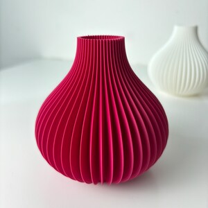 May include: A magenta vase with a ribbed design, shaped like an onion. A second white vase is visible in the background. These decorative vases are suitable for home decor and measure approximately 15 cm in height.