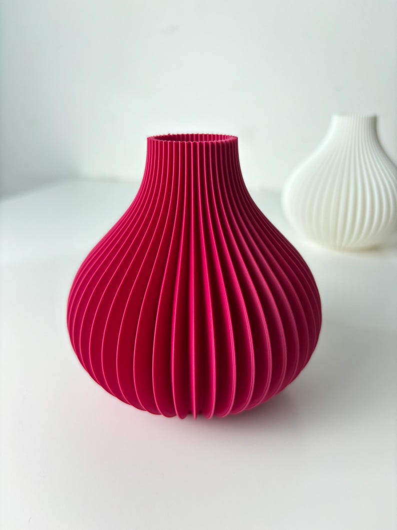 May include: A magenta vase with a ribbed design, shaped like an onion. A second white vase is visible in the background. These decorative vases are suitable for home decor and measure approximately 15 cm in height.