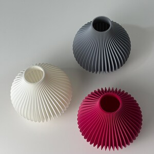 May include: Three decorative vases in white, grey, and magenta. Each vase has a round body with a fluted, fan-like design and a narrow opening at the top. The vases are arranged on a white surface.