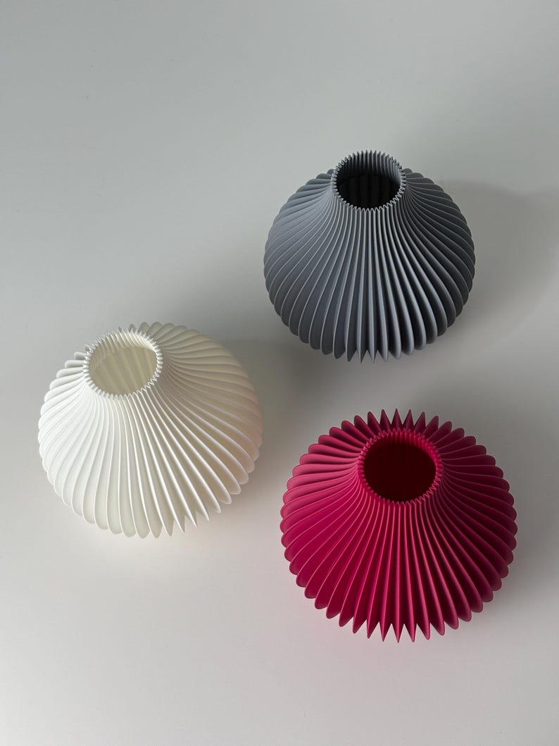May include: Three decorative vases in white, grey, and magenta. Each vase has a round body with a fluted, fan-like design and a narrow opening at the top. The vases are arranged on a white surface.