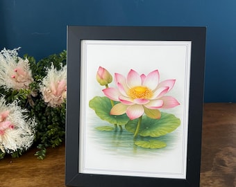 Op de afbeelding: Een ingelijste print met een gedetailleerde illustratie van een bloeiende roze lotusbloem met een geel hart en een gesloten lotusknop, tegen een achtergrond van groene waterleliebladeren en kalm water, in een zwarte rechthoekige lijst.