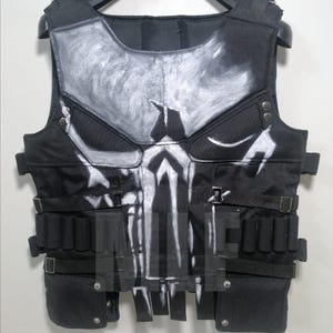 Può includere: Gilet tattico nero con un disegno a teschio bianco sul davanti. Il gilet presenta più tasche, cinghie e fibbie. È appeso a una gruccia nera.