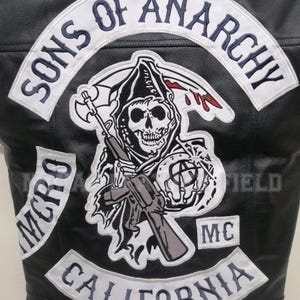 May include: Black leather vest featuring embroidered patches. The back displays a large patch with the words "SONS OF ANARCHY" above a grim reaper skull graphic holding a scythe and gun. Additional patches read "MC" and "CALIFORNIA".