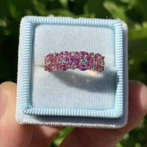 Può includere: Un anello d'oro con pietre preziose rosa e piccole pietre trasparenti, presentato in un astuccio di velluto blu chiaro. L'anello presenta una fila di pietre rosa ovali e pietre trasparenti più piccole.