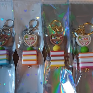 May include: Keyrings with a heart-shaped bookshelf design, available in silver and gold. Each keyring has a stack of colourful books and a round accent. The keyrings are packaged in clear plastic.