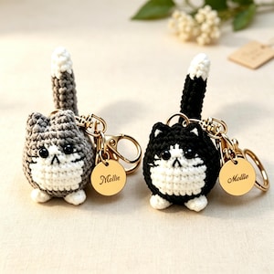 Könnte beinhalten: Zwei gehäkelte Katzen-Schlüsselanhänger, einer in Grau-Weiß und einer in Schwarz-Weiß, jeweils mit aufrechtem Schwanz, schwarzen Perlenaugen und einem goldfarbenen Metallring mit einem runden Anhänger, der den Namen Mollie in Schreibschrift trägt.