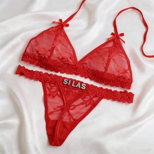May include: A red lace lingerie set, including a triangle bra and thong, with the name "SILAS" spelled out in silver rhinestones. The set features delicate floral patterns and small bows.