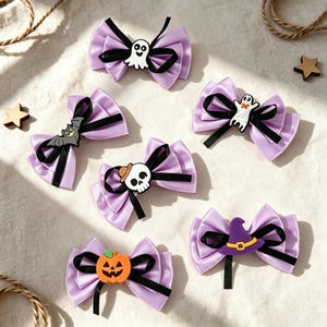 Könnte beinhalten: Sechs lavendelfarbene Haarschleifen aus Satin mit schwarzen Bandakzenten und verschiedenen Halloween-Motiven, darunter ein weißes Gespenst, eine graue Fledermaus, ein Totenkopf mit Hut, ein Kürbis, ein Geist mit Fliege und ein violetter Hexenhut.