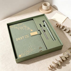 May include: A sage green 2027 daily plan notebook set in a matching box. The notebook cover features gold star patterns, a gold rectangular clasp, and the text 2027 DAILY PLAN and Iris. The set includes a matching sage green pen and a metal ink refill.