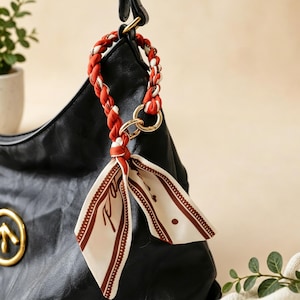 May include: A black handbag featuring a gold circular logo and a decorative accessory attached to the handle. The accessory consists of a braided orange and white fabric strap connected to a gold ring, with a dangling cream-coloured silk scarf.