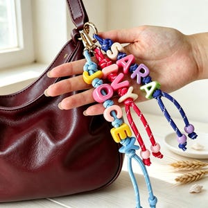 Può includere: Un set di tre portachiavi in corda colorata attaccati a una borsa in pelle rosso scuro. Ogni portachiavi presenta perline con lettere in plastica che compongono i nomi SARA, JOCEY e IVY, con piccole perline rotonde alle estremità dei cordini.